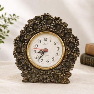 Puede incluir: Un reloj de cuarzo decorativo con un diseño de girasol de color bronce. La esfera del reloj es blanca con números negros y ribete dorado. Las manecillas del reloj son rojas. Es un elemento decorativo para el hogar.