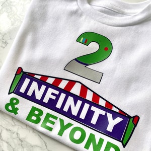 May include: White t-shirt with a green number 2 and a red, white, and blue striped banner that says "INFINITY & BEYOND" in green and purple letters.