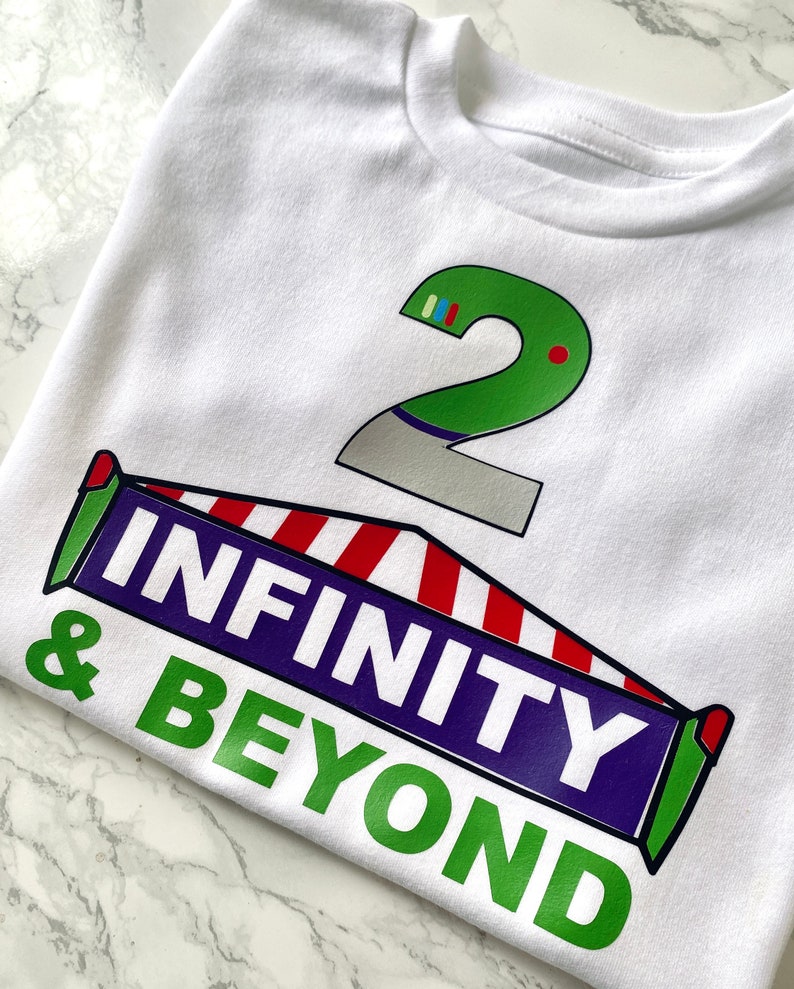 May include: White t-shirt with a green number 2 and a red, white, and blue striped banner that says "INFINITY & BEYOND" in green and purple letters.