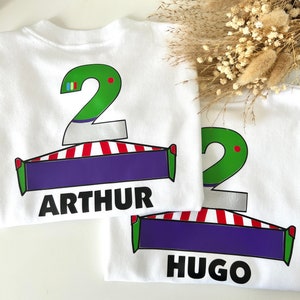 May include: Two white t-shirts with green number 2s, red and white striped designs, and purple banners. The top shirt has the name "ARTHUR" printed on the banner. The bottom shirt has the name "HUGO" printed on the banner.
