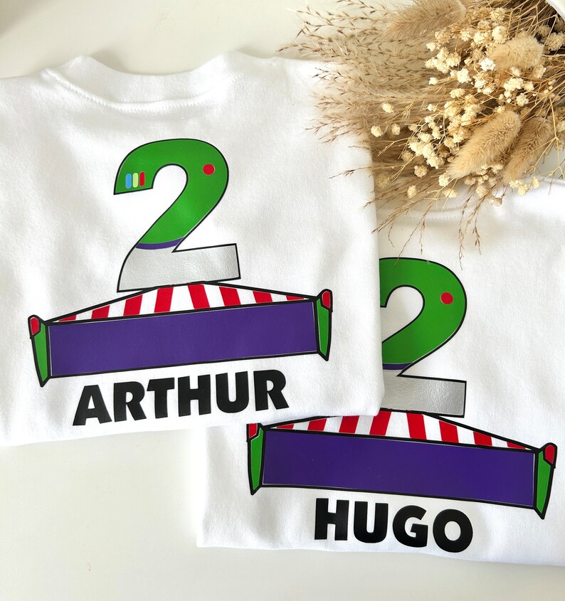 May include: Two white t-shirts with green number 2s, red and white striped designs, and purple banners. The top shirt has the name "ARTHUR" printed on the banner. The bottom shirt has the name "HUGO" printed on the banner.