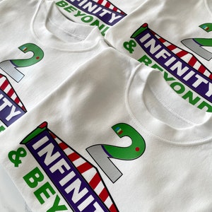 May include: Four white t-shirts with a green number 2 and the words "Infinity & Beyond" printed on them. The shirts are stacked on top of each other.
