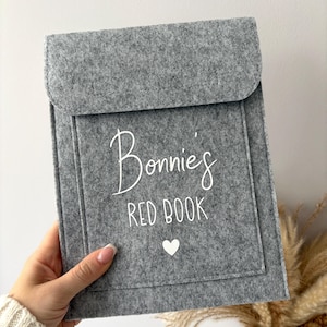 May include: A grey felt book cover with a flap and pocket. The cover is personalised with the text "Bonnie's RED BOOK" in white script and a small white heart. The book cover is held by a person.