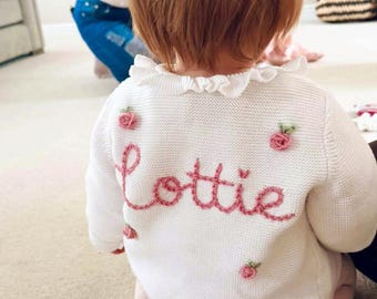 May include: A white long-sleeved knit cardigan featuring a ruffled collar and embroidered pink roses. The back of the garment displays the name Lottie stitched in pink cursive yarn, with small floral accents scattered across the white fabric.