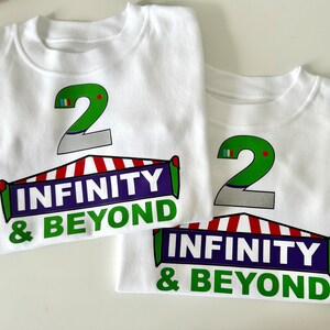 May include: Two white t-shirts with a green number 2 and the text "Infinity & Beyond" printed on them. The number 2 is on top of a red and white striped tent.