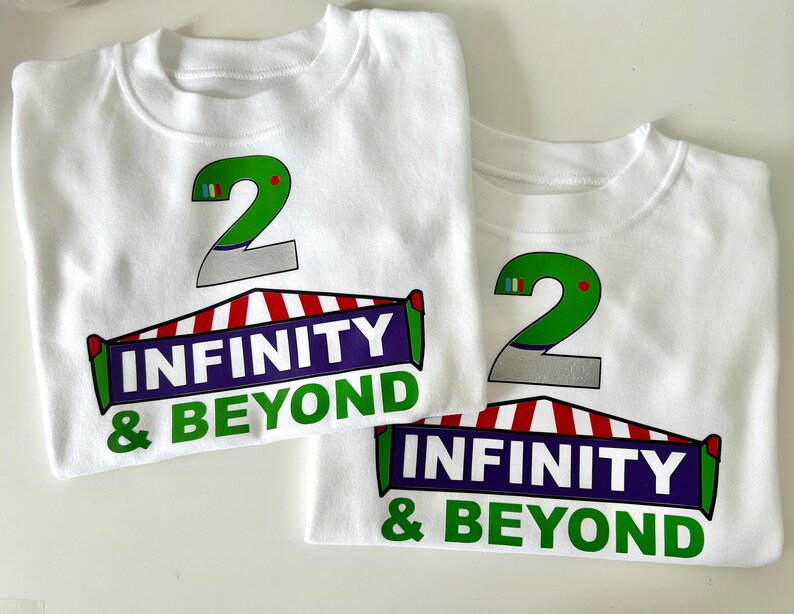 May include: Two white t-shirts with a green number 2 and the text "Infinity & Beyond" printed on them. The number 2 is on top of a red and white striped tent.