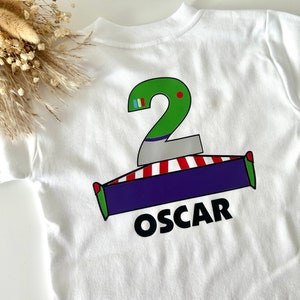 May include: White t-shirt with a green number 2, a red and white striped base, a purple rectangle, and the name "OSCAR" printed on the back.