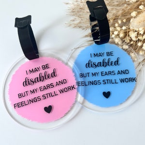 May include: Two round acrylic luggage tags with black straps. The pink tag says "I may be disabled but my ears and feelings still work" with a black heart. The blue tag says "I may be disabled but my ears and feelings still work" with a black heart.