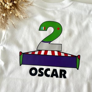 May include: White t-shirt with a green number two, red and white stripes, a purple rectangle, and the name "OSCAR" printed on the back.