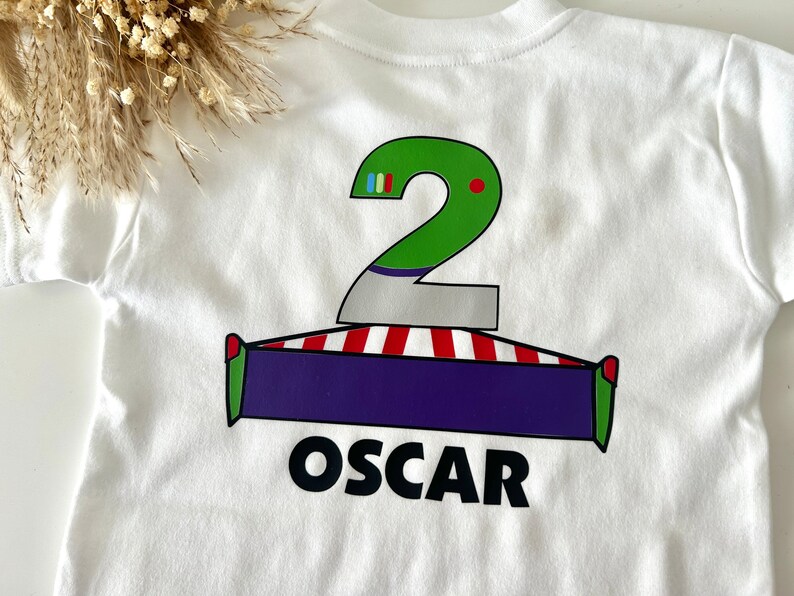 May include: White t-shirt with a green number two, red and white stripes, a purple rectangle, and the name "OSCAR" printed on the back.