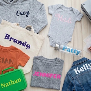 May include: A collection of personalized items including a gray hoodie with 'Greg', a cream tote bag with 'Brandy', an orange t-shirt with 'Brian', a green lunchbox with 'Nathan', a gray baby bodysuit with 'Heidi', a water bottle with 'Patty', a gray t-shirt with 'JENNIFER', and a blue denim jacket with 'Kelly'.