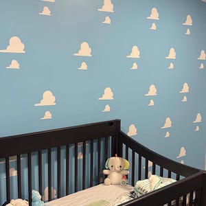 May include: A dark brown wooden crib with vertical slats containing a plush elephant toy and a white and gray striped blanket. The wall behind the crib is painted light blue and features a repeating pattern of white, stylized, puffy clouds.