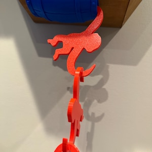 May include: A bright blue barrel-shaped object mounted to a wooden base, with a chain of three interlocking red monkey-shaped figures hanging from it. Each red monkey figure features a hook-like tail and curved arms, creating a vertical link.
