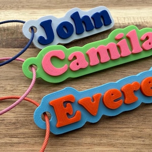 May include: Three custom luggage tags with raised lettering. One white tag with dark blue text reading John, one light green tag with pink text reading Camila, and one light blue tag with orange text reading Everett. Each tag has a colored wire loop.