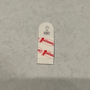 May include: A white Command brand adhesive strip with red text that reads "Command" and a small arrow pointing upwards. The strip is designed for hanging items on walls.