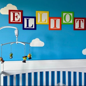 May include: A white crib with a blue and white cloud wall decal and a mobile hanging above it. The mobile has four small toys hanging from it. The wall also has the name "ELLIOT" spelled out in colorful blocks.