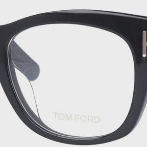 May include: Black Tom Ford spectacles with a thick, rounded frame. The glasses feature a silver accent on the temples and the Tom Ford logo on the lens. The frame is glossy and the lenses are clear.