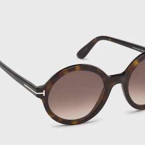 Może przedstawiać: Okrągłe okulary przeciwsłoneczne z wzorem w kolorze żółwia i brązowymi soczewkami gradientowymi. Okulary mają ciemnobrązowe zauszniki i złoty akcent na zausznikach. Soczewki są w kolorze jasnobrązowym.