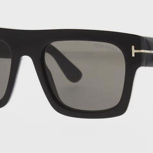 May include: Black sunglasses with a square frame. The sunglasses have gray lenses and gold-colored accents on the temples. The brand name "TOM FORD" is visible on the lens.