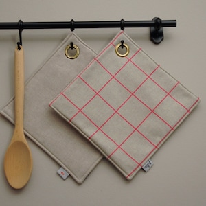 Op de afbeelding: Twee beige keukenonderzetters met roze ruitjespatronen die aan een zwarte metalen rek hangen. De onderzetters hebben messing oogjes en zijn gelabeld "Faded".