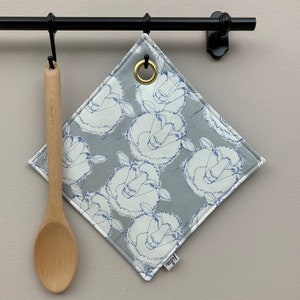 Könnte beinhalten: Ein grauer und weißer Topflappen mit einem blauen Schafdesign. Der Topflappen hat eine Messingöse und hängt an einem schwarzen Metallgestell.
