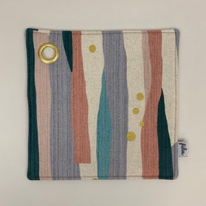 Op de afbeelding: Een vierkante, beige ovenwant met een kleurrijk abstract ontwerp in tinten roze, blauw, groen en grijs. De ovenwant heeft een gouden metalen oogje in de linkerbovenhoek en een klein wit label met het woord "fides" in zwarte letters in de rechterbenedenhoek.