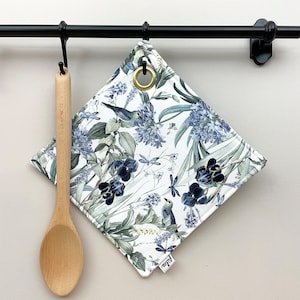 Könnte beinhalten: Ein weißer Baumwoll-Topflappen mit einem Blumenmuster mit blauen Blumen, grünen Blättern, Libellen und Vögeln. Der Topflappen hat einen goldenen Metallring und das Wort "Frida" ist in der rechten unteren Ecke aufgedruckt.