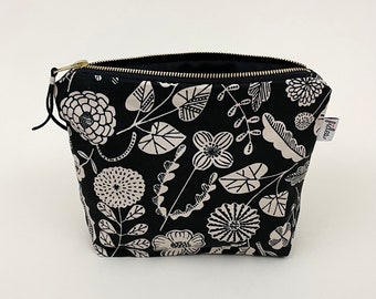 Op de afbeelding: Zwarte toilettas met rits en een wit bloemenpatroon. De tas heeft een gouden rits en een klein wit label met het woord "fiber" erop.