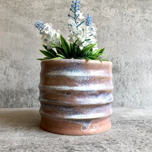 Pode incluir: Um vaso de cerâmica artesanal com um design texturizado e canelado em tons de castanho, branco e roxo. O vaso contém flores artificiais brancas e azuis com folhas verdes.