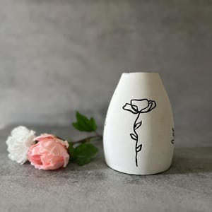 Op de afbeelding: Witte keramische vaas met een zwarte lijntekening van een bloem. De vaas staat op een grijs oppervlak.