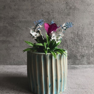 Puede incluir: Un macetero de cerámica azul y blanco con textura acanalada, lleno de una pequeña disposición de flores artificiales, incluyendo flores blancas y moradas y ramitas azules.