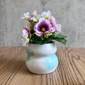 Peut inclure: Un petit pot en céramique blanche avec un glaçage bleu clair. Le pot a une forme arrondie et bulbeuse et est rempli d'un bouquet de fleurs artificielles. Les fleurs sont blanches et violettes, avec quelques feuilles vertes.