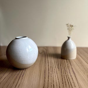 Op de afbeelding: Twee keramische vazen op een houten oppervlak. De grotere vaas is bolvormig, wit met gespikkelde details en een bruine basis. De kleinere vaas is van een vergelijkbare kleur, met een smalle hals en bevat gedroogde bloemen. De achtergrond is neutraal beige.