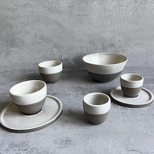 Può includere: Un set di stoviglie in ceramica fatto a mano composto da una ciotola grande, tre tazze piccole e due piattini rotondi. Ogni pezzo presenta un design bicolore con uno smalto bianco lucido sulla parte superiore e una finitura grigio scuro opaca.
