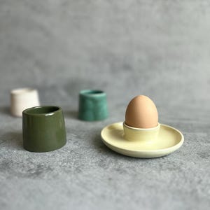 Puede incluir: Un huevero de cerámica amarillo claro con un huevo marrón y una taza de cerámica verde. En el fondo hay otras dos tazas de cerámica de diferentes colores. Los objetos están sobre una superficie gris.