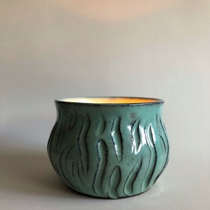 Op de afbeelding: Een kleine, teal-kleurige keramische plantenpot met een getextureerd, bladvormig ontwerp. De pot heeft een afgeronde bodem en een licht uitlopende rand.