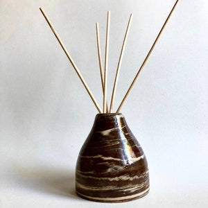 Può includere: Un flacone diffusore in ceramica marrone e bianco con bastoncini di legno che sporgono dalla parte superiore.