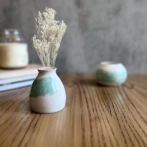 Può includere: Un piccolo vaso in ceramica bianca con una glassa blu chiaro, che contiene un mazzo di fiori bianchi secchi. Il vaso è appoggiato su un tavolo di legno.