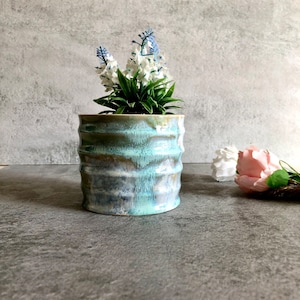 Puede incluir: Jardinera cilíndrica de cerámica con esmalte turquesa texturizado. La jardinera contiene un grupo de flores artificiales blancas y azules. Una flor rosa y blanca está en el fondo.