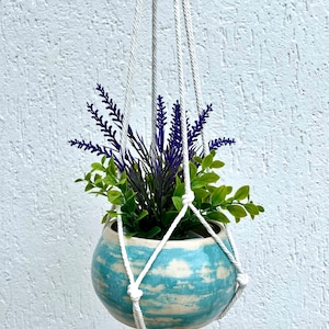 Può includere: Un portavasi in macramè bianco con un vaso in ceramica blu e bianco. Il vaso è pieno di una pianta verde e fiori viola.
