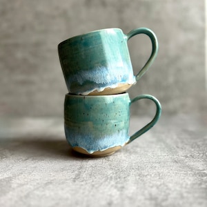 Può includere: Due tazze in ceramica impilate con una glassa blu chiaro e verde. Le tazze hanno una finitura macchiata e un bordo bianco.