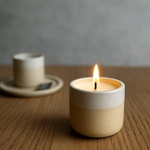 Puede incluir: Una vela encendida en un recipiente de cerámica de dos tonos. La vela tiene una parte superior blanca y una base amarilla. Una pequeña taza y platillo de cerámica a juego están en el fondo. La llama de la vela es brillante.