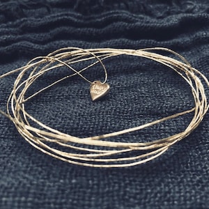 Peut inclure: Un collier avec un pendentif en forme de cœur en or gravé des mots "Always Made". Le collier est sur un fond en tissu bleu.