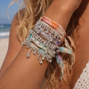 May include: A collection of colorful fabric bracelets adorned with sparkling, dangling rhinestones. The bracelets are tied around an arm, with a beach scene in the background. The fabric has a tie-dye effect in shades of pink, orange, purple, and blue.