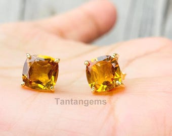 May include: A pair of square-cut, golden-yellow gemstone stud earrings with gold prongs. The earrings are displayed on a light background. The text "Tantangems" is visible.