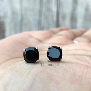 May include: A pair of stud earrings featuring square-cut black gemstones set in silver-toned metal prongs. The text Tantangems is visible between the two earrings. Each earring has a faceted surface that reflects light across the dark stone.