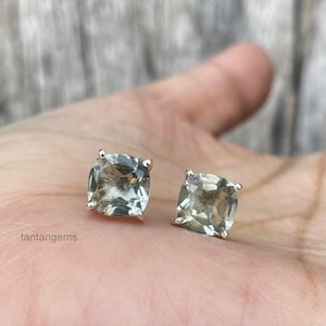 May include: A pair of square-cut, light green gemstone stud earrings with silver posts. The earrings are displayed on a light skin tone background. The earrings are a jewelry item.