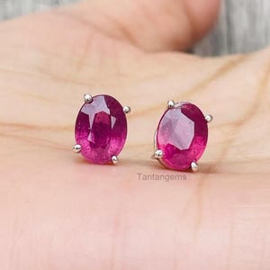 May include: A pair of oval-cut, deep pinkish-red ruby stud earrings, each secured in a four-prong silver-toned metal setting. The faceted gemstones exhibit a vibrant, translucent quality with visible internal clarity and light reflection.