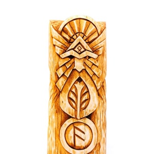 Puede incluir: Un tótem de madera con tallas intrincadas, incluyendo un símbolo solar, una hoja y una runa. El tótem está pintado en tonos marrones y beige.