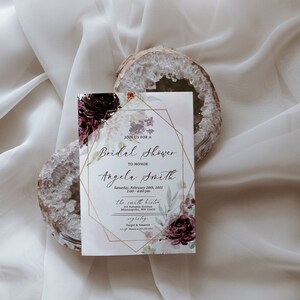 May include: A white bridal shower invitation with a gold geometric frame and floral design. The invitation reads "Join us for a Bridal Shower to honour Angela Smith" and includes the date, time, and location of the event.
